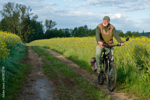 An active senior cycles early in the morning on a sunny field path, surrounded by brightly blooming yellow rapeseed fields, enjoying the peace and quiet of the morning hours.