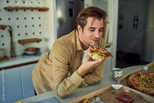 A smiling man leaning on a kitchen counter and enjoying a slice of pizza