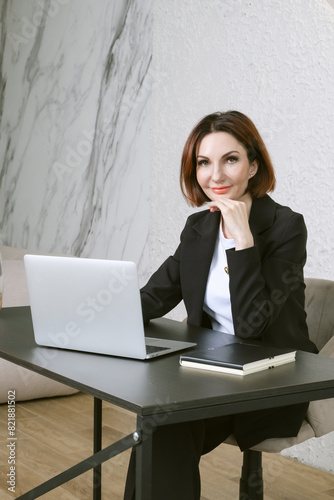 Portrait of business woman in a black business suit at desk in your office. Business portrait. Business woman is sitting in front of laptop screen
