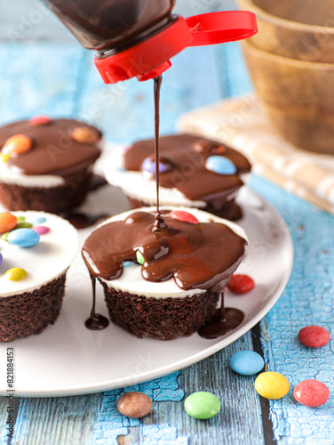 Chocolate sauce is poured from a bottle onto a plate with cupcakes. The cupcakes have white icing and are topped with colorful candy-coated chocolates. In the background, a wooden bowl is partially.