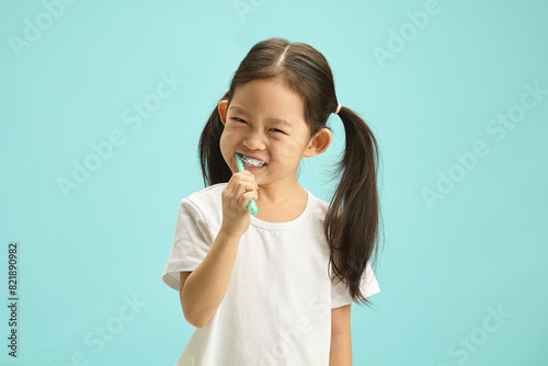 Face of Japanese child girl cleaning mouth with a brush for morning routine and oral self care. Happy asian ethnicity kid using toothbrush standing over blue isolated background with free copy space.