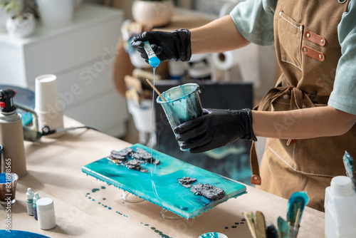 Female artist pours epoxy resin and smears it with stick on wooden board. Craftswoman using liquid art technique, creating picture of sea and waves, closeup view. Art deco, master class on resin art.  photo