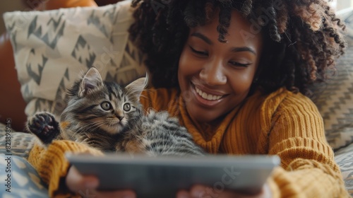 A woman smiling while using a tablet and relaxing on a couch with her cat, enjoying quality time in a cozy home setting.
