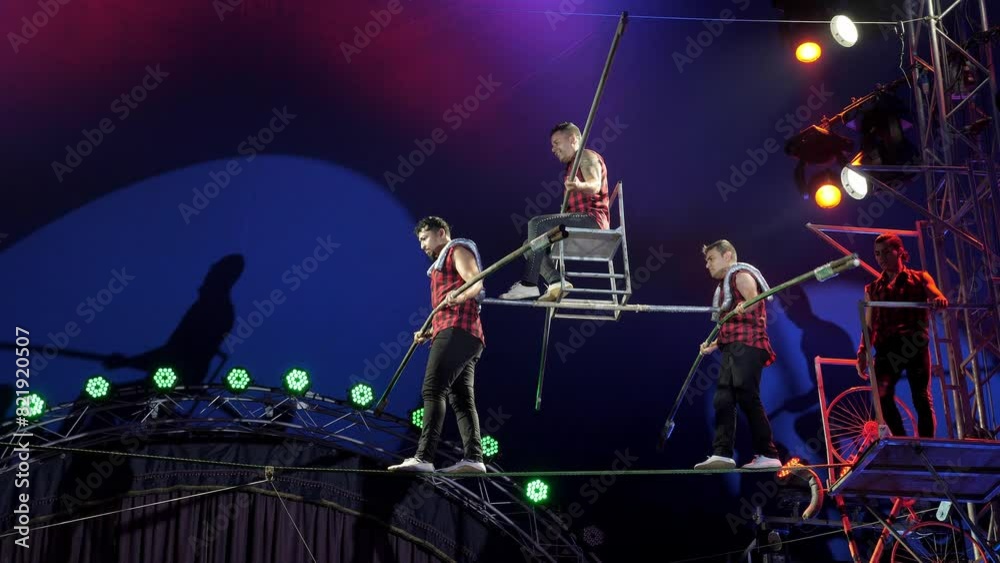 Circus acrobats perform on a tightrope in a circus, two acrobats hold a third on their shoulders. Concert lighting with multi colored spotlights. Three tightrope walkers perform a difficult trick.