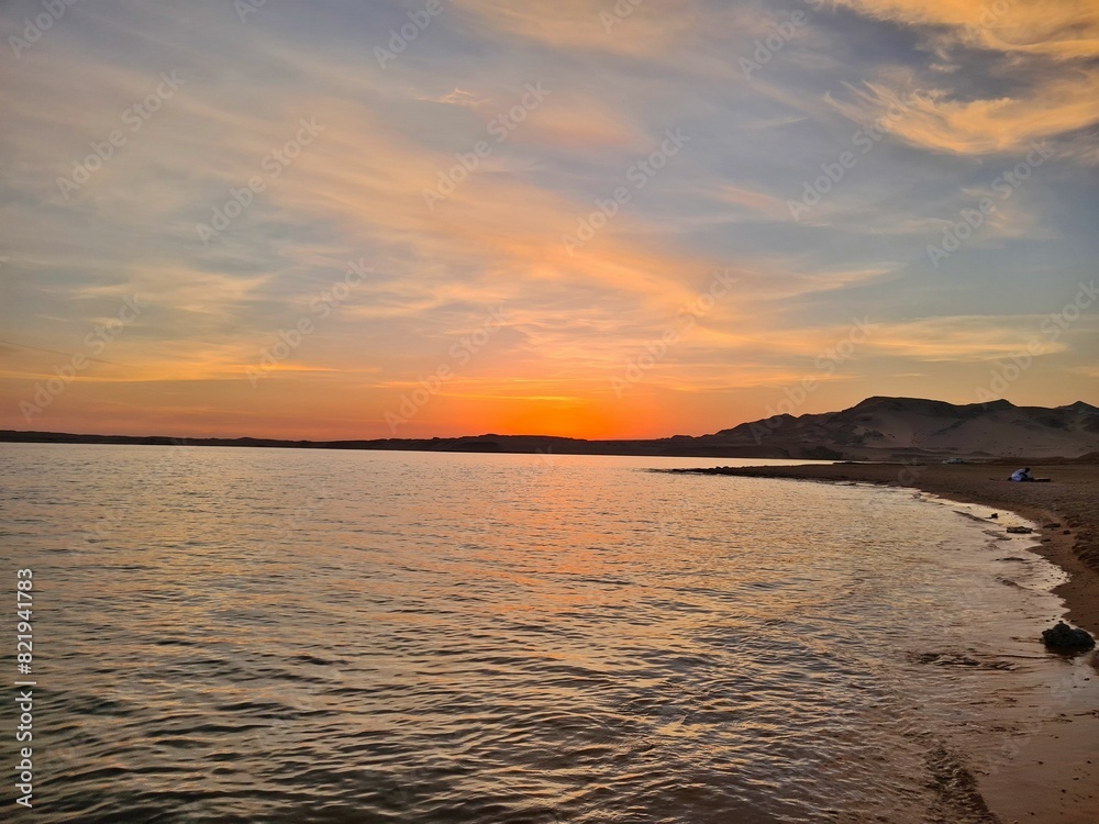 Naklejka premium Orange and pink sunrise peeking over the horizon of a tranquil beach, reflecting on the calm water
