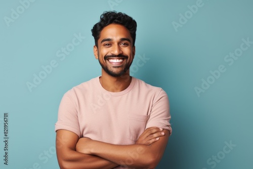 Portrait of a grinning indian man in his 20s with arms crossed over solid pastel color wall