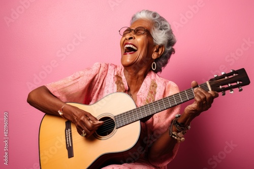 Portrait of a cheerful indian woman in her 80s playing the guitar while standing against solid pastel color wall