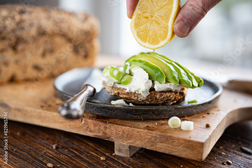 Fresh avocado slices on grain bread with cream cheese. Hand from above with lemon. Vegetarian food on wooden cutting board. Close-up in front of a bright window.