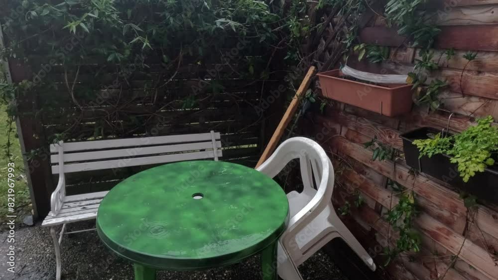 Raindrops pouring on a round green table and chairs by the wall and plant trellis in the backyard