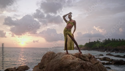 Godlike gender fluid person of color in open dress, jewelry stands on cliff peak at scenic ocean sunrise. Pride extravagant fashion model in luxury outfit posing like goddess in tropics. Olymp concept photo