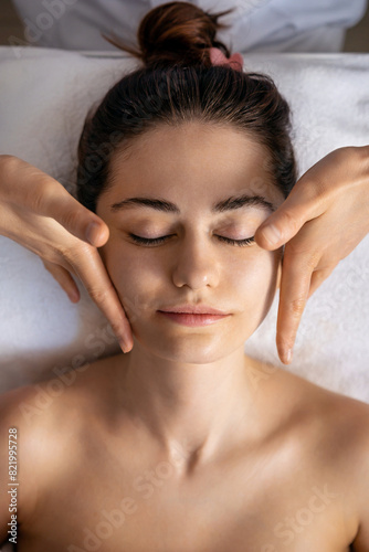 From above view of a calm female closing eyes and enjoying face massage during session in spa salon. Facial beauty treatment, body care, wellness
