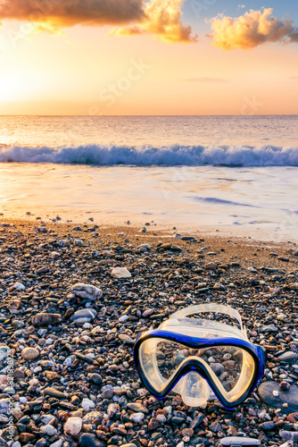 ocean cloudy morning landscape with diving mask on sea coast on foreground, sand of a beach, blue sea with surf and waves and cloudy sunset or sunrise on background