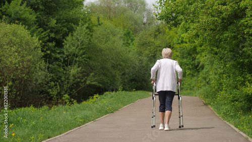 Walking along a path bordered by trees and grass, an elderly woman uses a walker. © StockMediaProduction
