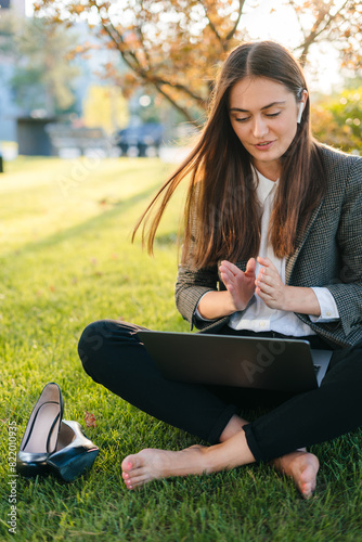 Young caucasian woman sitting at park lawn having a video conference call on her laptop. Female sitting on grassy lawn having a video call on laptop.