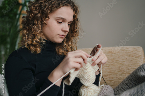 Young woman with curly hair knitting wool product sitting on sofa at home. Knitting product from white woolen thread.