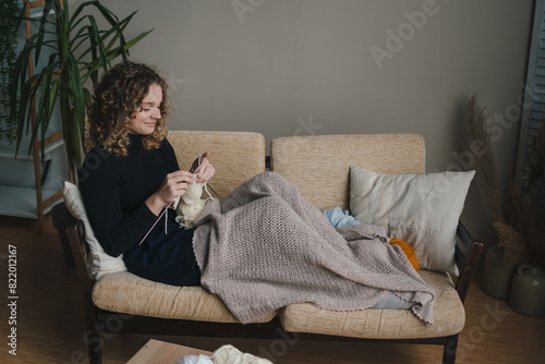 Young caucasian woman knitting handmade piece while sitting on couch at home and enjoying cozy hobby.