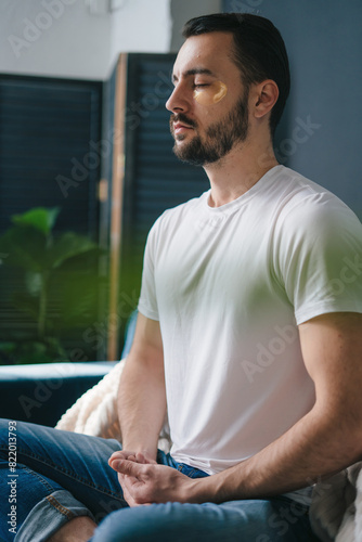 Focused adult man meditating in lotus pose breathing deep and slowly sitting on comfortable coach relaxing at home. Relaxing after a working day, meditating.