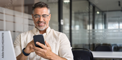 Banner with middle-age Hispanic business manager ceo using cell phone mobile app. Smiling Latin or Indian mature man businessman holding smartphone in office, working as trader on pc, with copy space