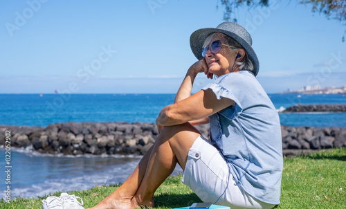 Smiling senior woman in straw hat sitting barefoot in meadow face the sea listening to music, relaxed senior lady enjoying free time in summer vacation or retirement