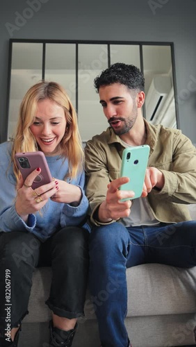 Vertical. Female holding a credit card, shopping online and her husband pointing her smart phone. Adult couple searching a sale on a cellphone social media app sitting on a couch at home living room