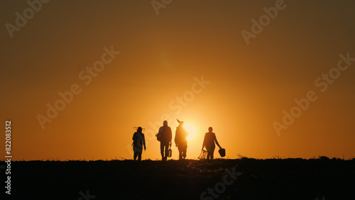 With the sun setting behind them, a family of farmers carries agricultural implements as they walk across a field.