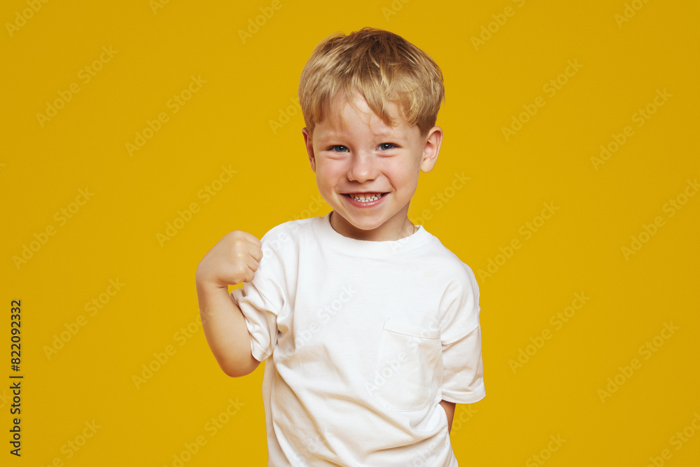 Portrait of little cute shy kid boy with blonde hair wearing white tshirt, standing and looking at camera against yellow background. Studio shoot. Portrait of little cute shy kid boy with blonde hair wearing white tshirt, standing and looking at camera against yellow background. Studio shoot.