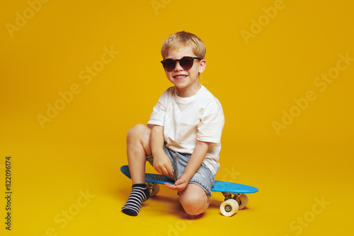 Cool little kid boy wearing casual outfit and sunglasses smiling while sitting on skateboard against yellow studio backdrop