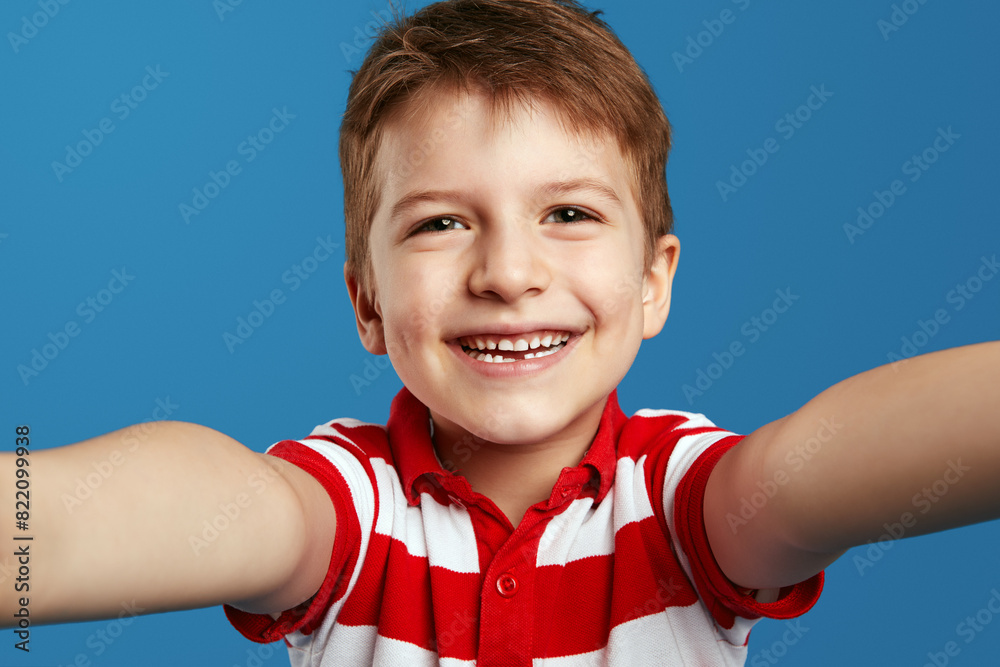 Funny child boy in red striped polo shirt looking at camera in amazement and taking selfie against blue background Funny child boy in red striped polo shirt looking at camera in amazement and taking selfie against blue background