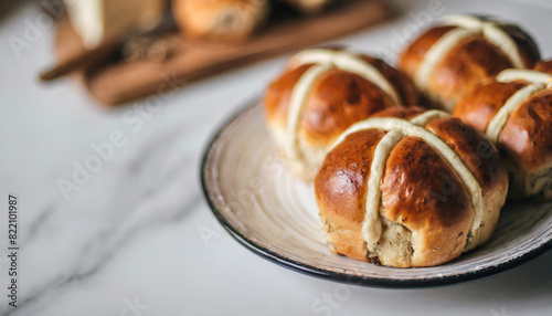 hot cross buns on a plate on a white table, blurred kitchen background. Freshly baked, lightly glazed, with iconic cross on top, evoking tradition and festive warmth