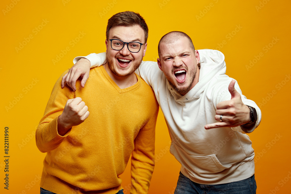 Two crazy excited unshaven guys screaming while showing horn sign, standing isolated over yellow background. True friendship concept Two crazy excited unshaven guys screaming while showing horn sign, standing isolated over yellow background. True friendship concept
