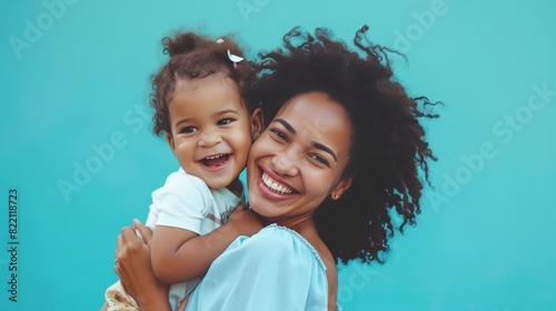 A young African American mother with curly hair joyfully holding her toddler daughter against a blue background.