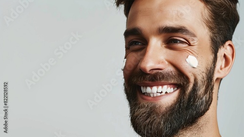 A cheerful man with a wellgroomed beard and facial cream dots on his cheek The light gray background highlights his radiant skin and happy demeanor photo