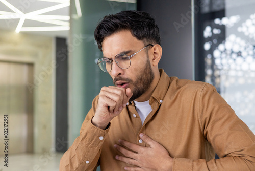 Young man coughing in modern office environment, hand on chest