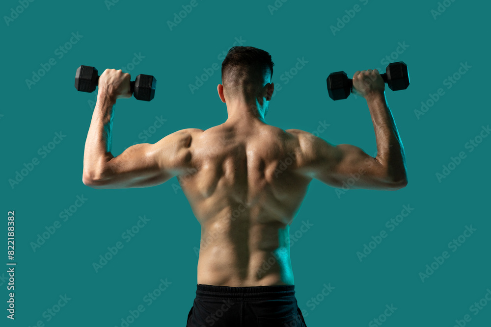 A man is seen from the back performing a shoulder press with dumbbells in each hand. His muscular back is highlighted against a solid teal background A man is seen from the back performing a shoulder press with dumbbells in each hand. His muscular back is highlighted against a solid teal background