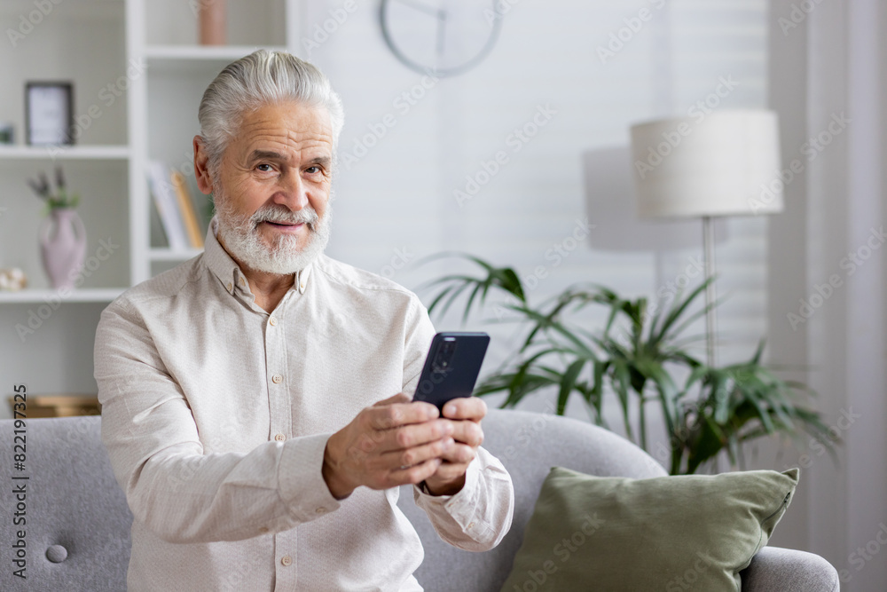 Portrait of an older gray-haired man in a white shirt sitting on the couch at home, holding a mobile phone and smiling at the camera Portrait of an older gray-haired man in a white shirt sitting on the couch at home, holding a mobile phone and smiling at the camera