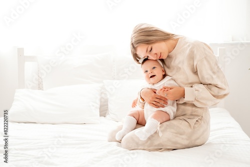 a mother with a newborn baby in her arms at home on a white background of a bed or window, a space for text, a young mother gently kisses and hugs her baby, maternal love
