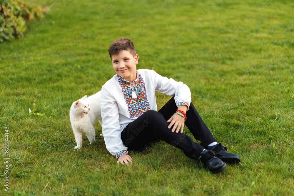 Young teenager in an embroidered shirt posing while sitting on the lawn playing with a white cat