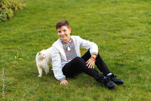 Obraz na płótnie Young teenager in an embroidered shirt posing while sitting on the lawn playing with a white cat w salonie
