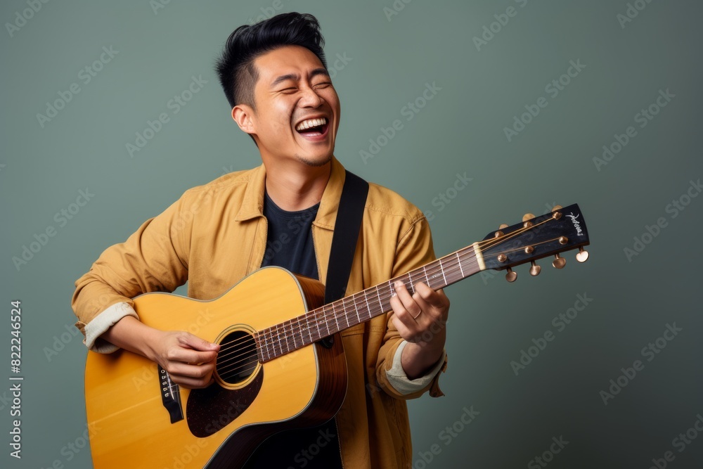 Portrait of a cheerful asian man in his 30s playing the guitar while standing against blank studio backdrop
