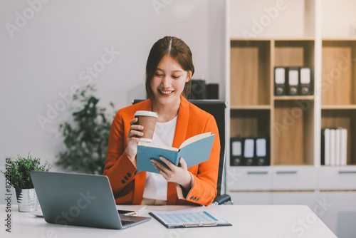 Beautiful smiling Asian businesswoman holding a coffee cup while taking notes at office.