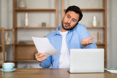 Fotografia A perplexed man is seated at a wooden desk, talking on phone, holding a document