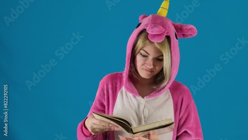 Girl in unicorn pajamas reads textbook standing on blue background. Female teenager shakes hands showing unwillingness to do hometask photo