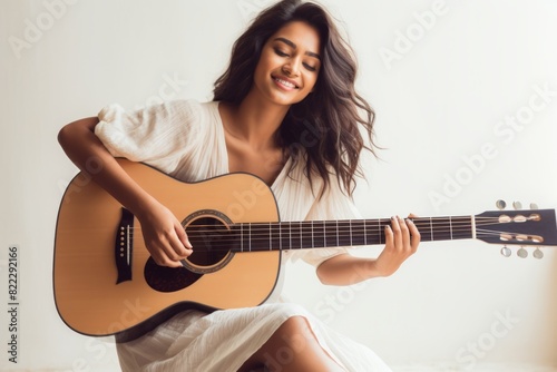 Portrait of a satisfied indian woman in her 20s playing the guitar isolated on light wood minimalistic setup