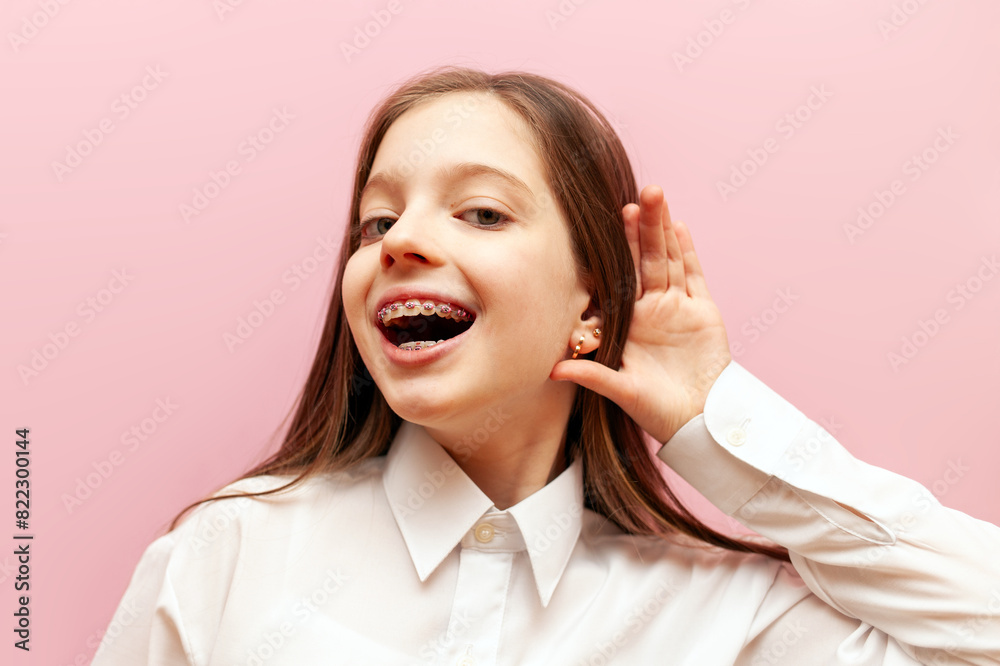 curious little teenage girl with braces eavesdrops on pink isolated background, child holds hand near ear and listens curious little teenage girl with braces eavesdrops on pink isolated background, child holds hand near ear and listens