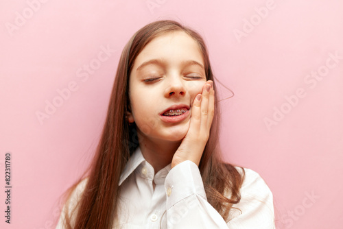 little teenage girl with dental braces suffers from toothache on pink isolated background, child holds cheek with hand and shows toothache