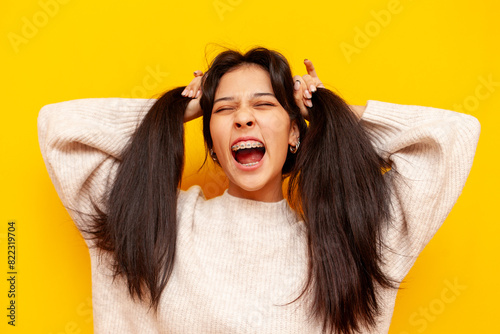 Young shocked asian woman with braces surprised with open mouth over yellow isolated background, excited korean girl screaming in panic and holding her hair in amazement