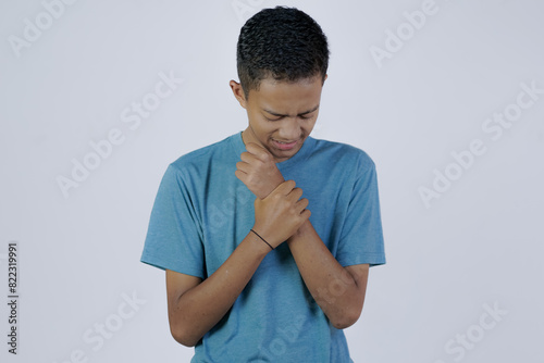 young Asian man in blue shirt moaning in pain feeling his sore wrist, joint disease, isolated white background. photo