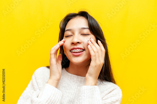 young Asian woman with braces smiles with closed eyes and touches the skin on her face with her hands on a yellow isolated background, Korean girl takes care of her skin and washes her face and