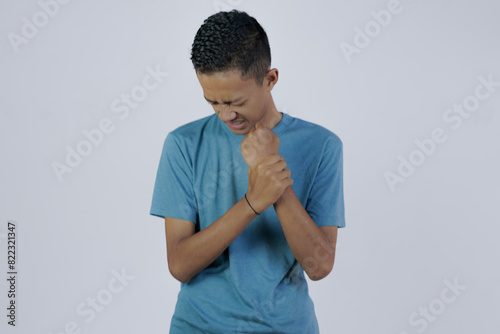 young Asian man in blue shirt moaning in pain feeling his sore wrist, joint disease, isolated white background. photo