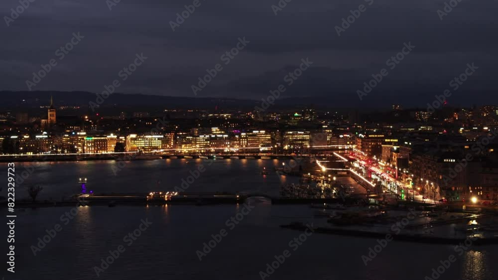 Aerial view of Geneva's waterfront at night, showcasing the city lights and their reflections on the water with Alps in the background Aerial view of Geneva's waterfront at night, showcasing the city lights and their reflections on the water with Alps in the background
